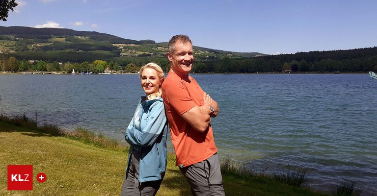Ein Mann und eine Frau stehen in der Nähe eines Sees. Sie lächeln beide und schauen zur Kamera. Der Mann hat die Arme verschränkt. Die Frau trägt eine Jacke und der Mann ein orangefarbenes T-Shirt.