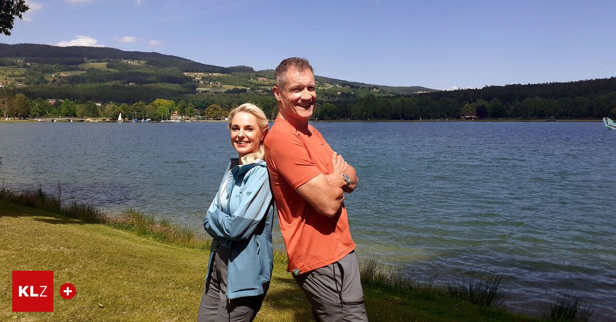 Ein Mann und eine Frau stehen in der Nähe eines Sees. Sie lächeln beide und schauen zur Kamera. Der Mann hat die Arme verschränkt. Die Frau trägt eine Jacke und der Mann ein orangefarbenes T-Shirt.