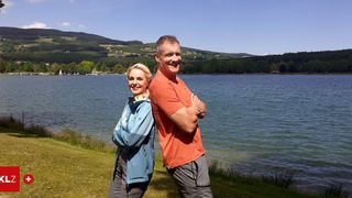 Ein Mann und eine Frau stehen in der Nähe eines Sees. Sie lächeln beide und schauen zur Kamera. Der Mann hat die Arme verschränkt. Die Frau trägt eine Jacke und der Mann ein orangefarbenes T-Shirt.