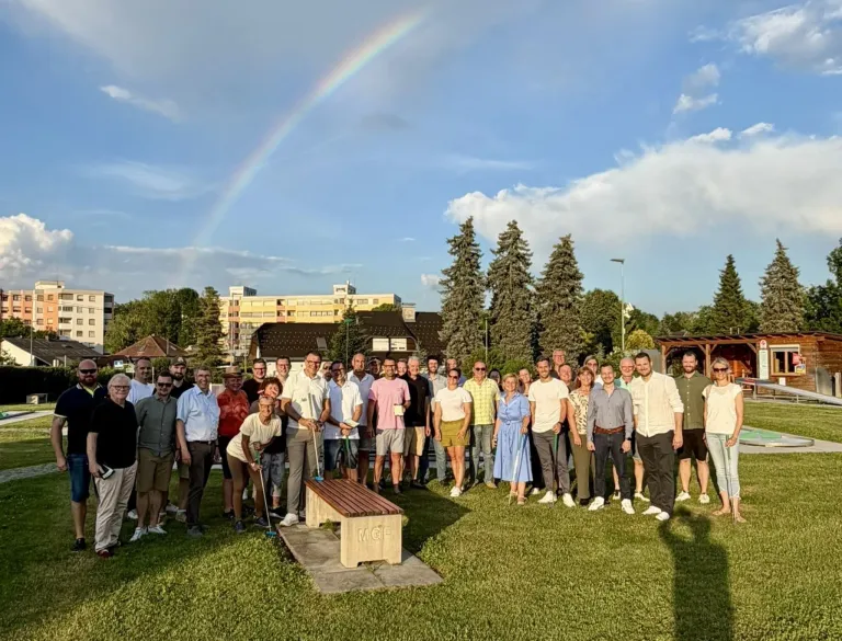 Eine Gruppe von Menschen steht in einem Park mit einem Regenbogen am Himmel und hält Golfschläger. Ein hölzerner Sitz mit den Buchstaben MGF steht im Vordergrund. Bäume und Gebäude sind im Hintergrund.