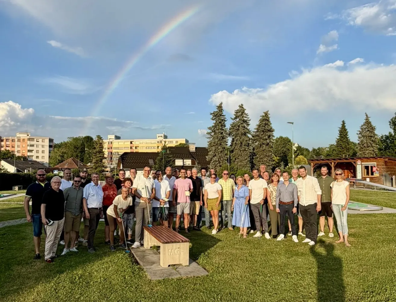 Eine Gruppe von Menschen steht in einem Park mit einem Regenbogen am Himmel und hält Golfschläger. Ein hölzerner Sitz mit den Buchstaben MGF steht im Vordergrund. Bäume und Gebäude sind im Hintergrund.