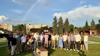 Eine Gruppe von Menschen steht in einem Park mit einem Regenbogen am Himmel und hält Golfschläger. Ein hölzerner Sitz mit den Buchstaben MGF steht im Vordergrund. Bäume und Gebäude sind im Hintergrund.