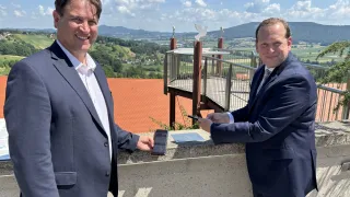 Zwei Männer in Anzügen stehen auf einem Balkon mit einem Telefon und blicken auf eine malerische Landschaft mit Bergen und einem Haus.