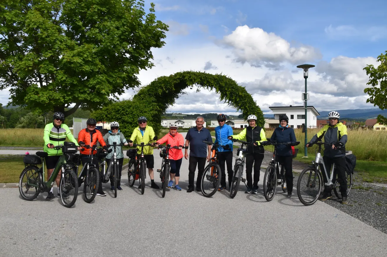 Eine Gruppe von Radfahrern in Sicherheitsausrüstung posiert für ein Foto unter einem grünen Torbogen. Dahinter befinden sich Häuser und ein Baum.