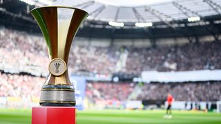 Ein goldener Pokal steht auf einer roten Plattform in einem Stadion mit verschwommenem Publikum. Ein Spieler steht auf dem Feld.