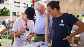 Drei Personen stehen an einem Tisch mit wissenschaftlichen Geräten. Eine Frau in einem weißen T-Shirt benutzt eine Pipette. Ein älterer Mann mit Brille schaut sie an. Ein anderer Mann in einem blauen T-Shirt beobachtet sie.