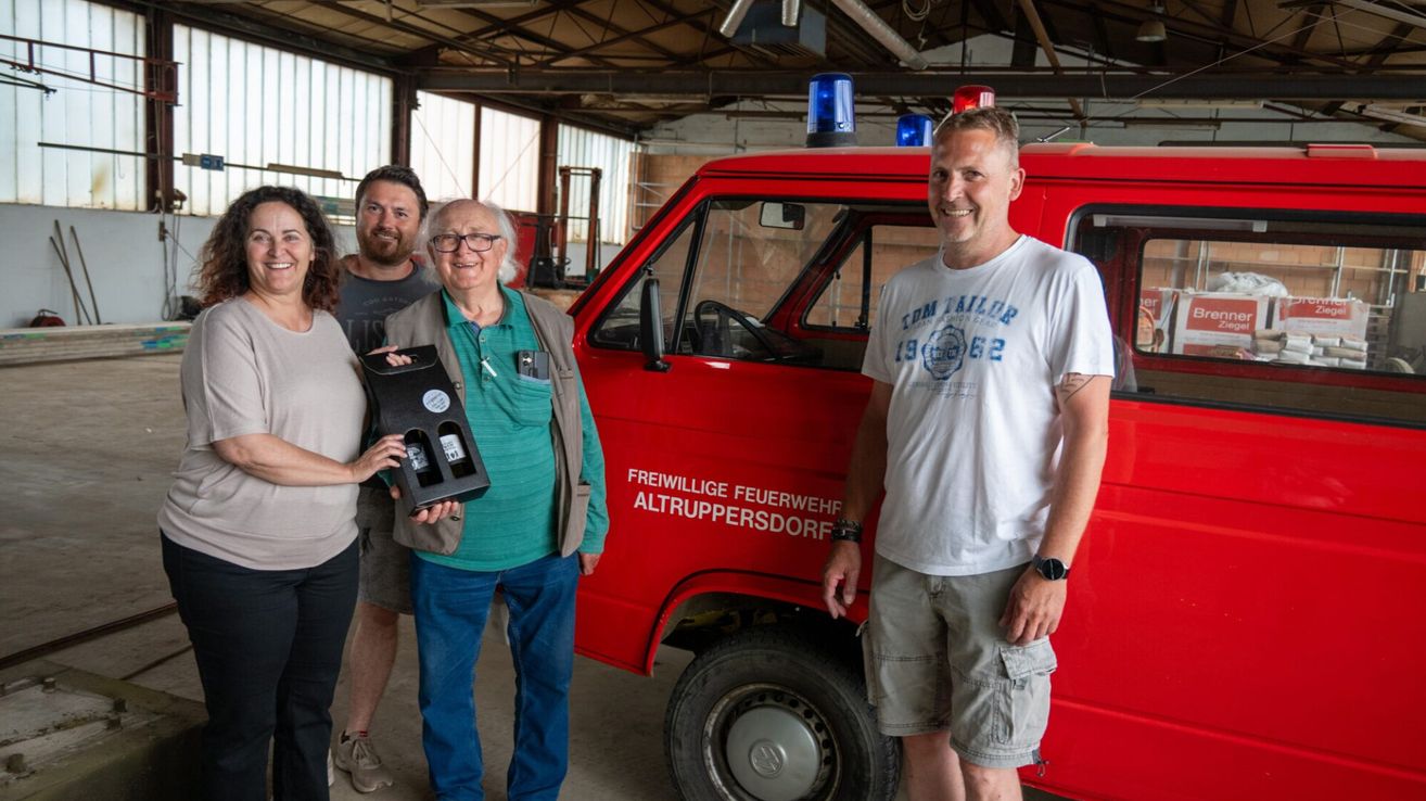 Vier Personen stehen vor einem roten Feuerwehrauto. Eine Person hält eine Weinbox und lächelt. Zwei Männer tragen Freizeitkleidung und eine Frau trägt einen hellen Pullover.