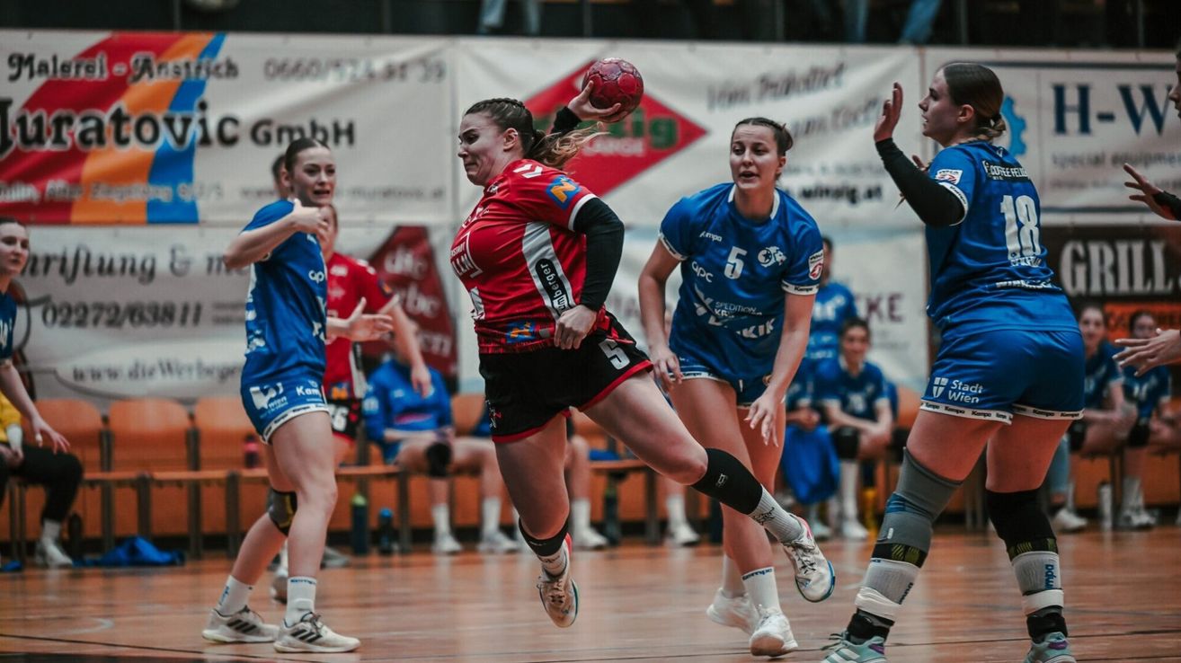 Frauenhandballspielerinnen in Aktion auf einem Spielfeld. Eine Spielerin in Rot bereitet sich auf den Ballwurf vor, während andere zusehen.
