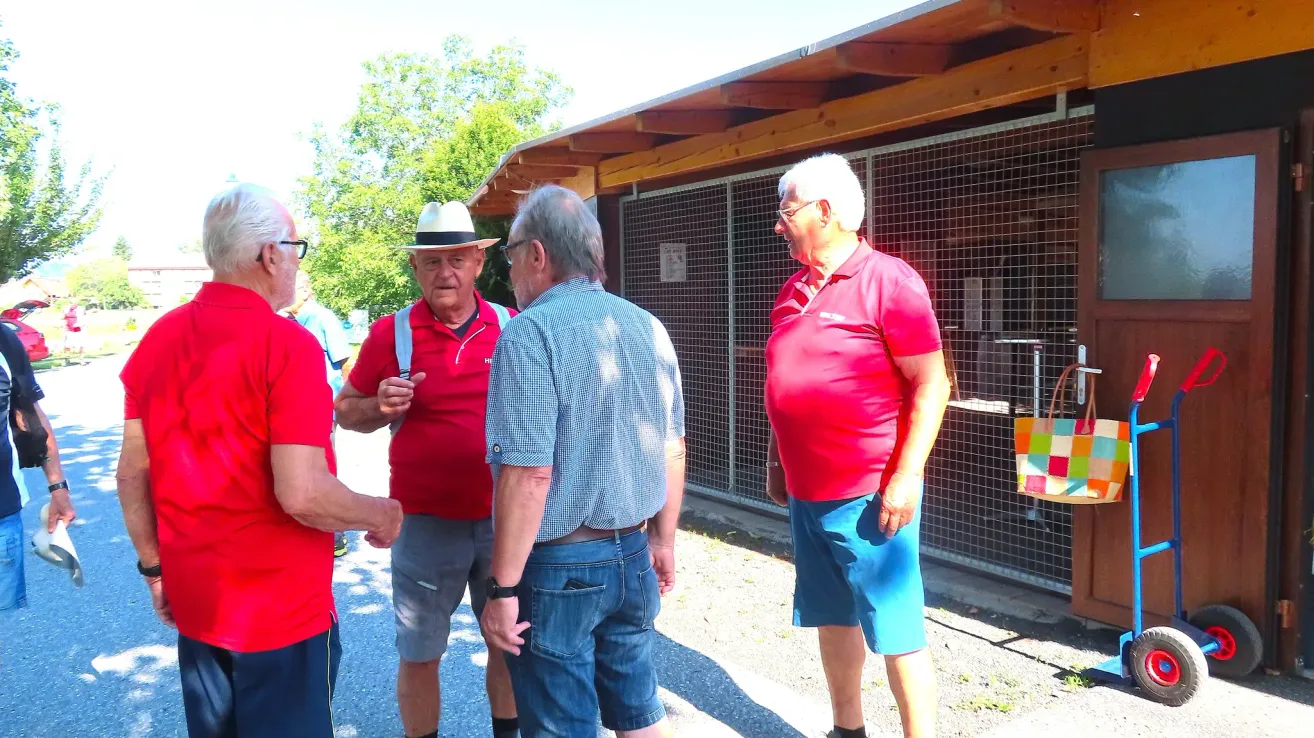 Drei Männer in Freizeitkleidung stehen vor einem Gebäude und unterhalten sich. Einer trägt einen Hut und Hosenträger, ein anderer trägt eine Brille. Hinter ihnen ist ein Zaun und Bäume zu sehen.