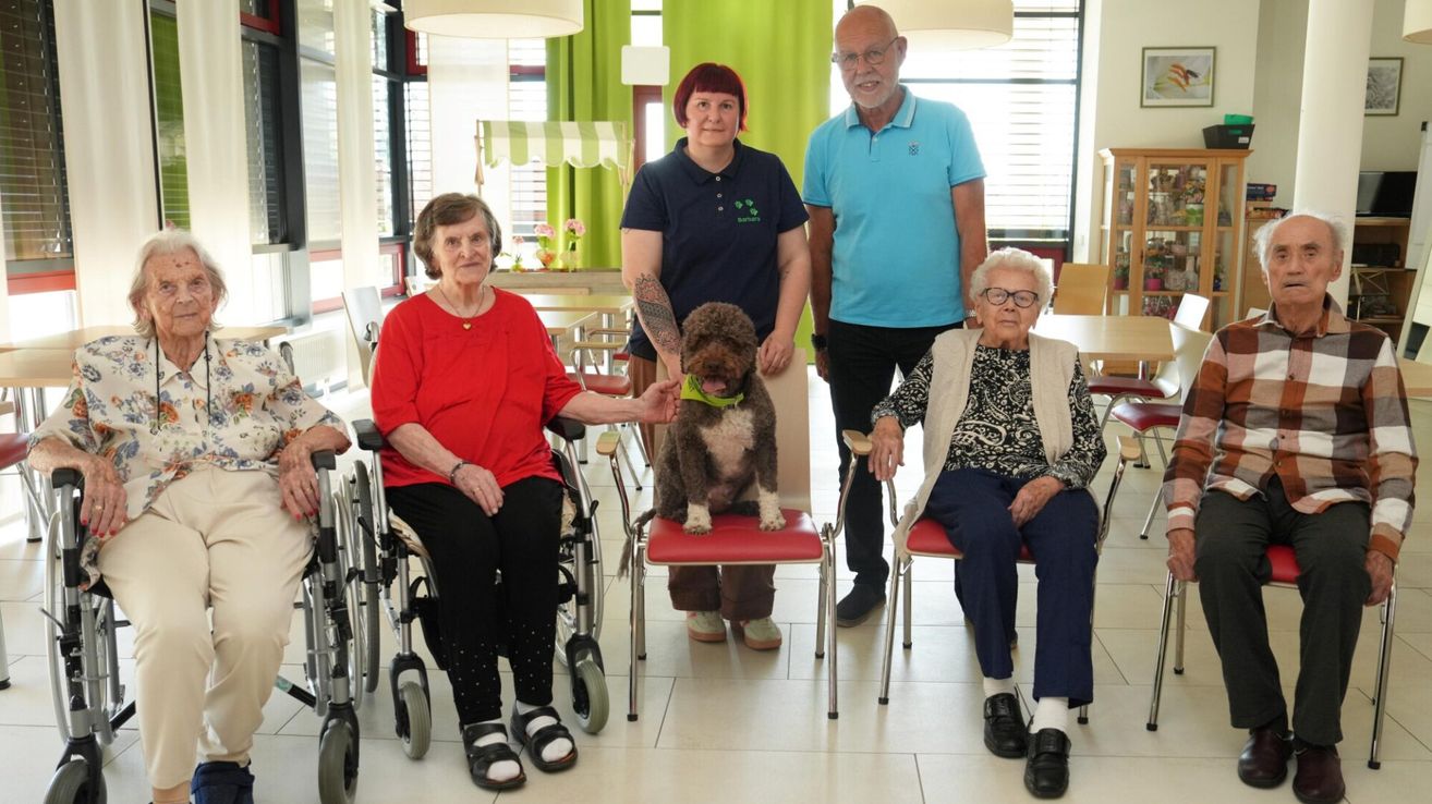 Vier ältere Menschen und ein Mann sind in einem Raum mit einem Hund auf einem Stuhl. Zwei ältere Frauen sitzen in Rollstühlen und eine weitere steht neben einem Stuhl. Der Mann trägt eine Brille und eine Uhr. Hinter ihnen stehen Tische und Stühle.