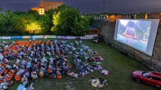 Ein Filmvorführung findet bei Nacht im Freien statt. Viele Menschen sitzen auf Stühlen und Decken auf dem Rasen und schauen auf die Leinwand. Ein Auto ist in der Nähe der Mauer geparkt.