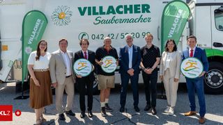 Eine Gruppe von Menschen steht vor einem weißen Lastwagen und hält grüne Schilder in der Hand, um 25 Jahre Villacher Saubermacher zu feiern. Sie tragen alle formelle Kleidung.
