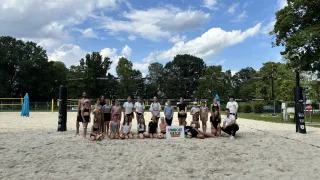 Eine Gruppe von Menschen, einschließlich Frauen und Männern, posiert für ein Foto auf einem sandigen Strand mit einem Volleyballnetz im Hintergrund.