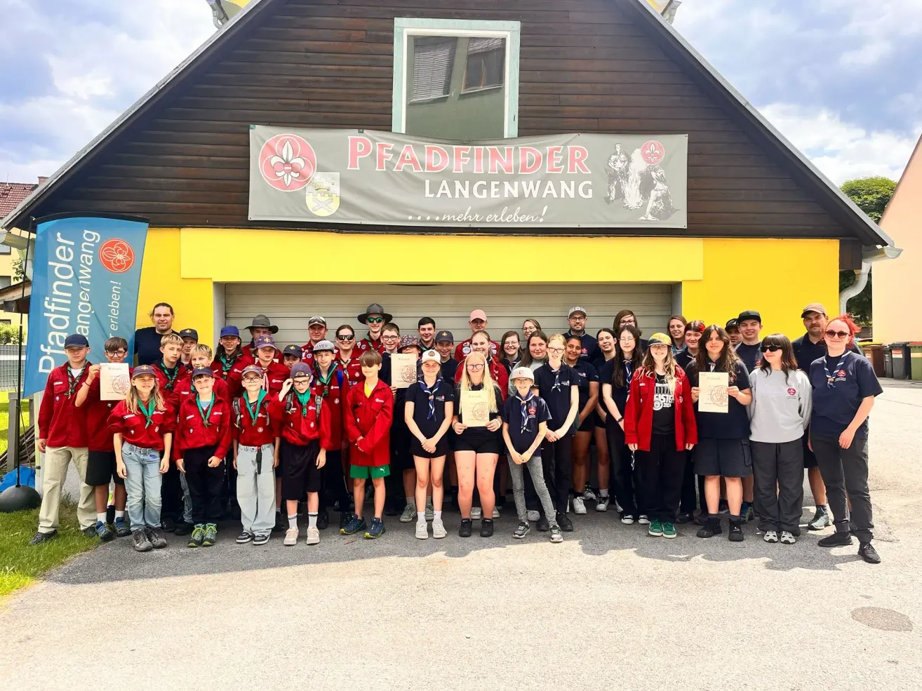 Eine Gruppe von Pfadfinder Langwang-Mitgliedern posiert für ein Foto vor einem Gebäude. Sie tragen alle rote und schwarze Uniformen und halten Urkunden.