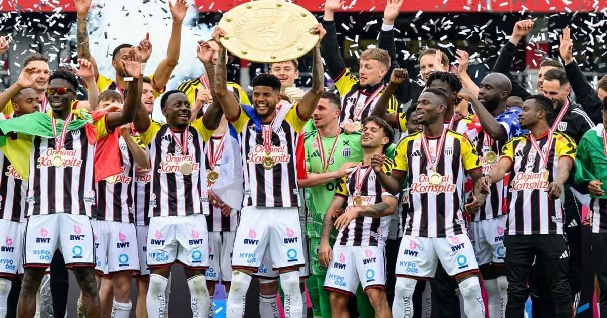 Fußballspieler feiern mit einer Trophäe und Medaillen vor einem jubelnden Publikum.