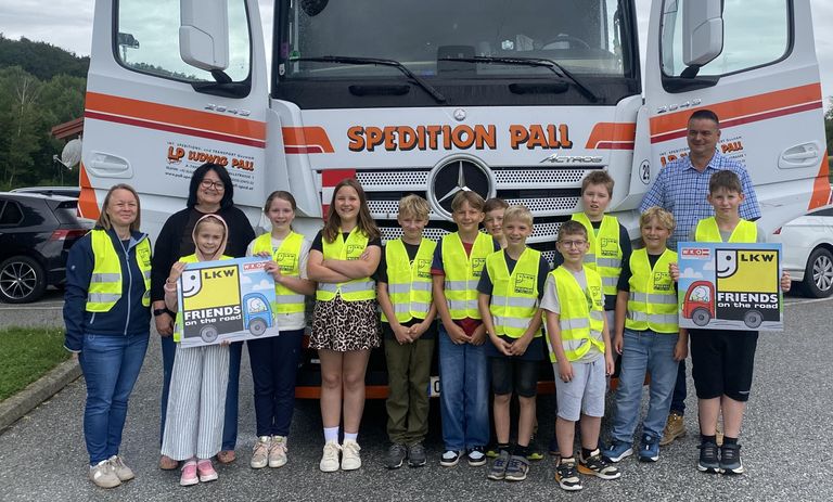 Eine Gruppe von Kindern und Erwachsenen steht vor einem mit 'SPEDITION PALL' beschrifteten LKW. Sie tragen gelbe Westen mit 'LKW'. Die Kinder halten ein Schild mit 'FREUNDE auf der Straße'. Der LKW ist weiß und orange.