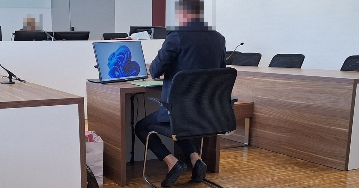 Eine Person sitzt an einem Schreibtisch in einem Büro, mit einem Laptop, der ein blaues abstraktes Design zeigt. Sie ist in formeller Kleidung und es steht ein Mikrofon auf dem Schreibtisch.
