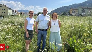 Drei Personen stehen in einer Wiese. Zwei Frauen und ein Mann, lächelnd, mit einem Hintergrund von Bergen und Häusern.