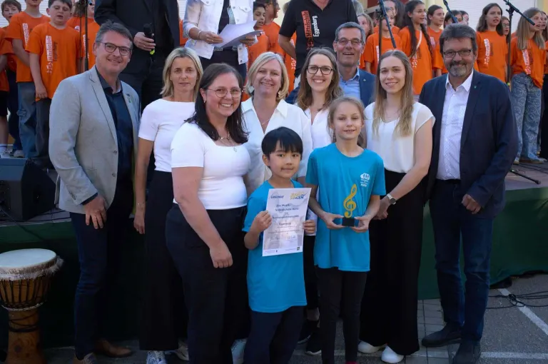 Eine Gruppe von Erwachsenen und Kindern posiert für ein Foto, wobei zwei Kinder Zertifikate halten. Sie lächeln und stehen in einer Reihe.