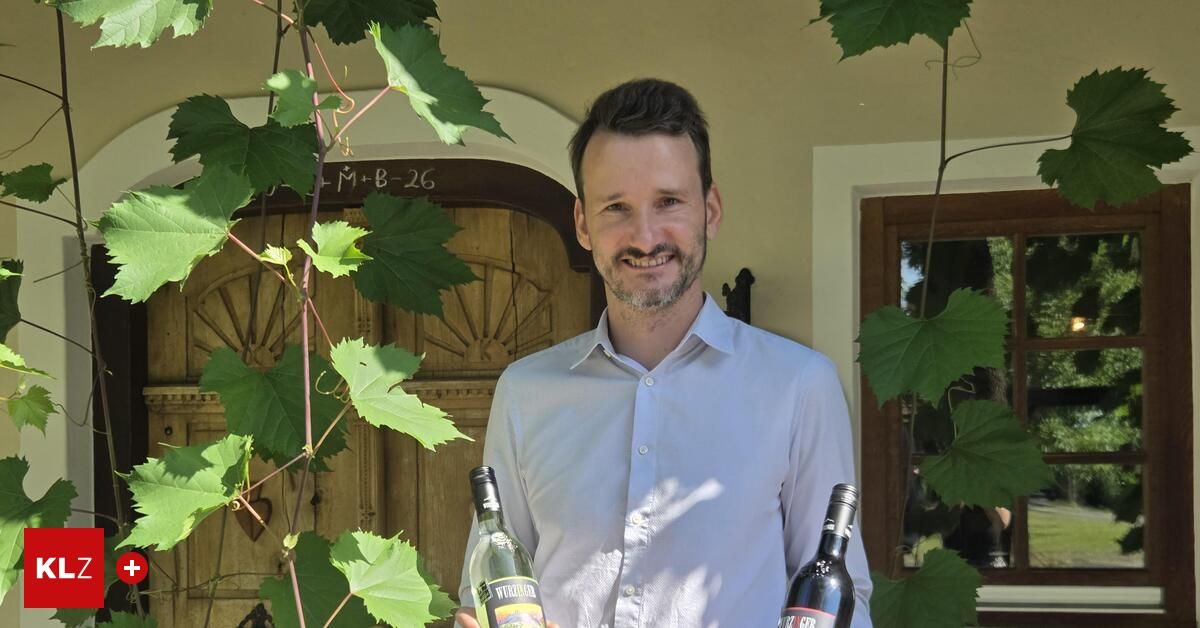 Ein Mann mit Bart und Schnurrbart lächelt, während er zwei Flaschen Wein vor einer Holztür mit einer Rebe hält.