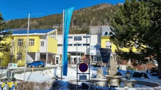 Ein Gebäude mit gelben und weißen Wänden hat Solarpanele und eine Flagge davor. Ein Auto ist auf der schneebedeckten Straße geparkt. In der Nähe gibt es ein Schild, Felsen und einen Baum. Ein Berg ist in der Ferne.