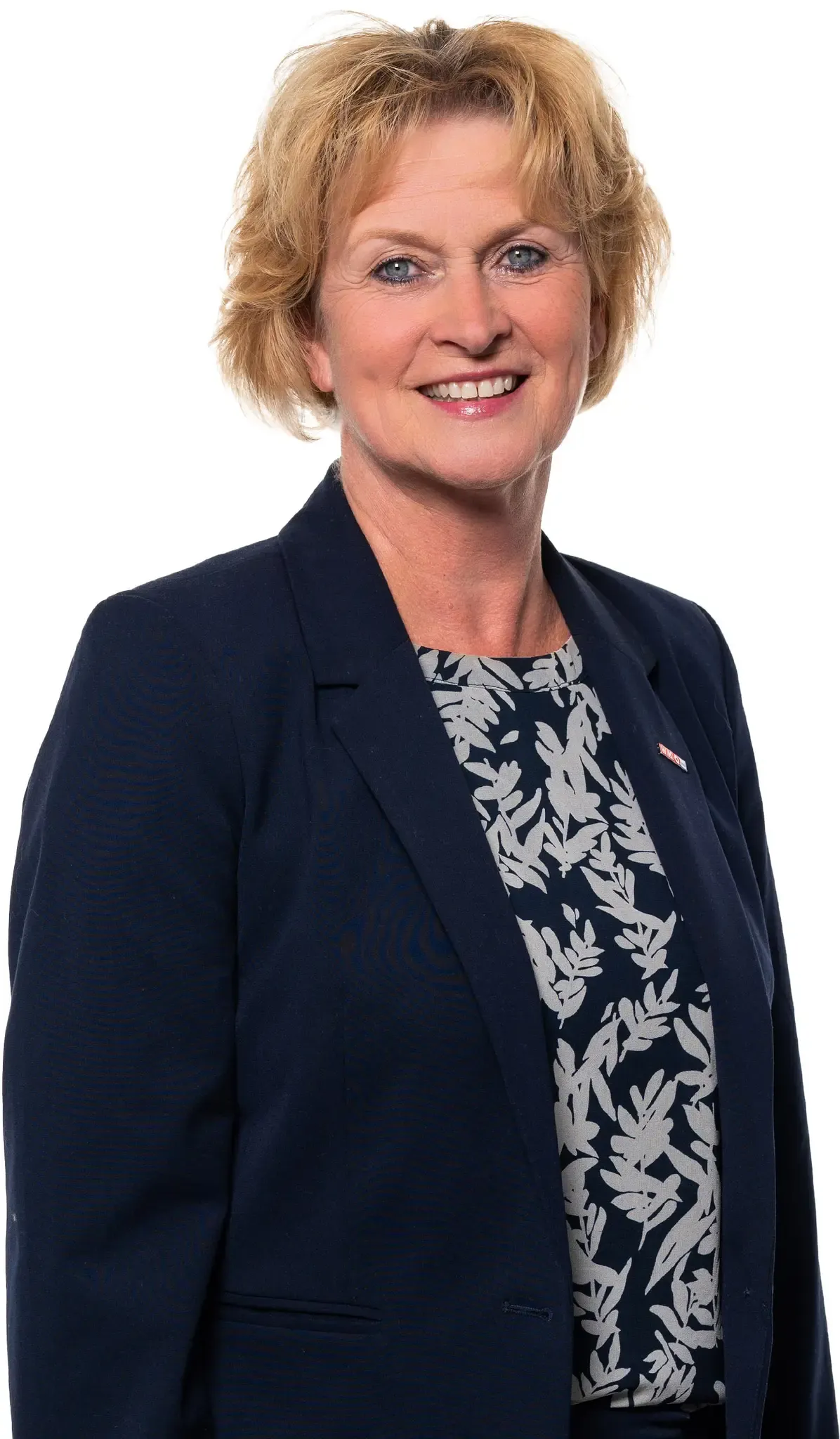 Eine Frau mit kurzen blonden Haaren, die einen marineblauen Blazer und ein weißes Blattdruck-Oberteil trägt, lächelt in die Kamera.