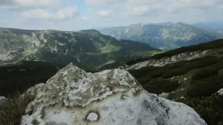 Ein Panoramablick von einem Berggipfel zeigt eine weite Landschaft mit üppigen grünen Tälern und rauen Bergketten unter einem bewölkten Himmel.