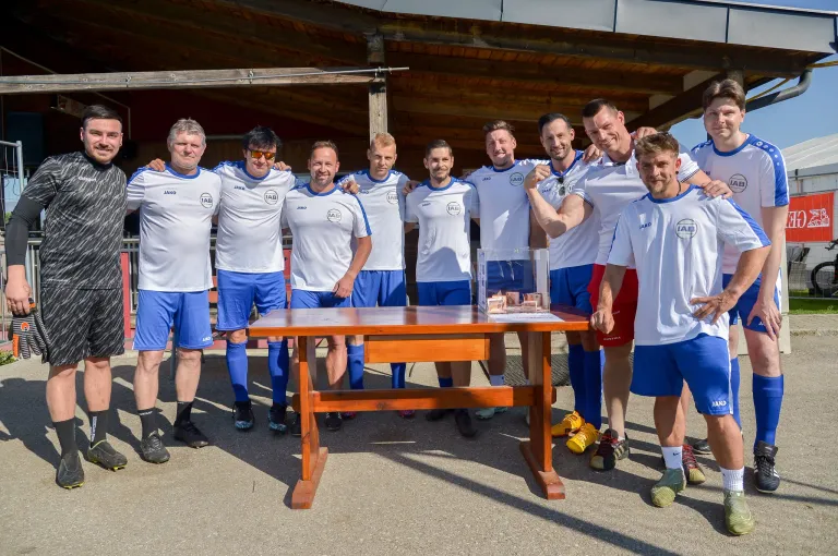 A group of male athletes wearing matching uniforms pose for a photo with a trophy on a wooden table in front of them.