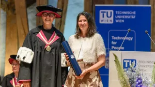 Eine Frau in einem Blumenrock steht neben einem Mann in akademischer Kleidung, beide lächeln. Sie stehen vor einem Banner für TU Wien.