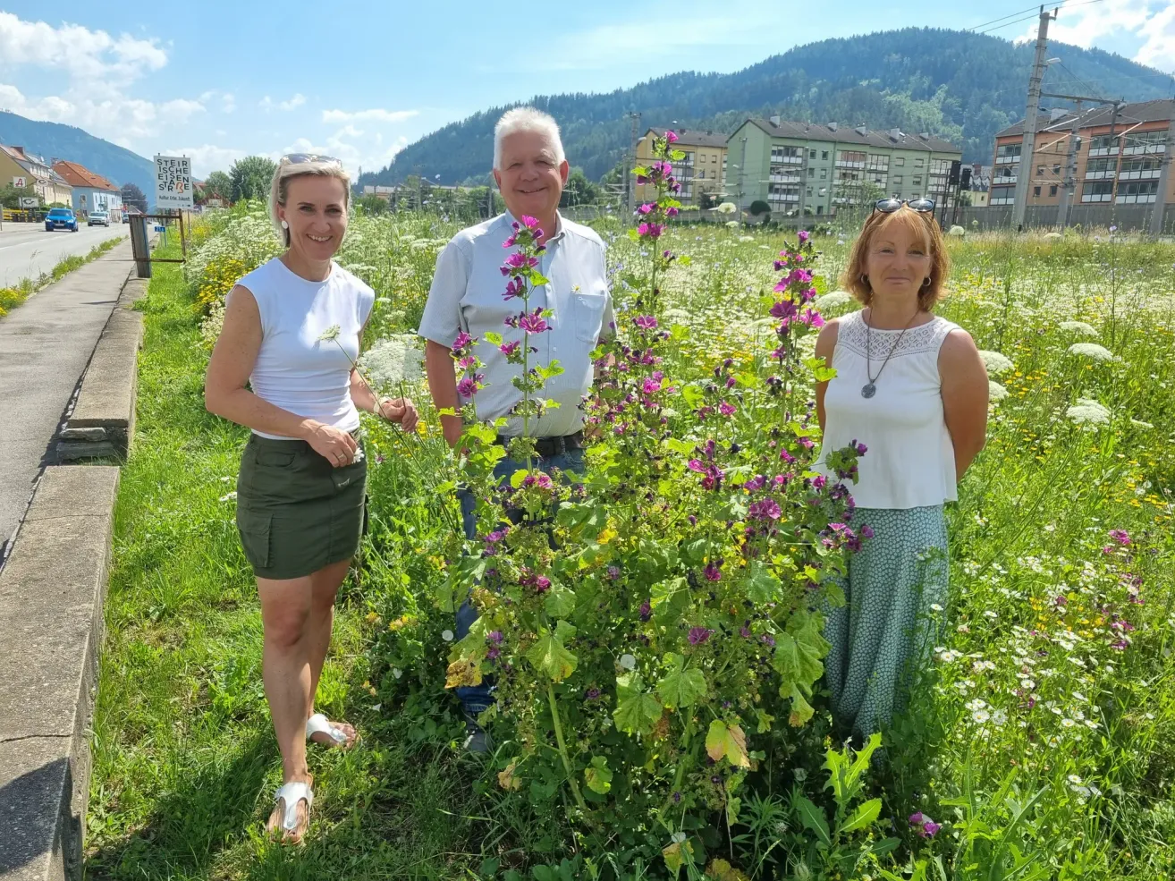 Drei Menschen stehen in einem Blumenfeld. Zwei Frauen und ein Mann, sie lächeln. Hinter ihnen sind Gebäude, Bäume und Berge.