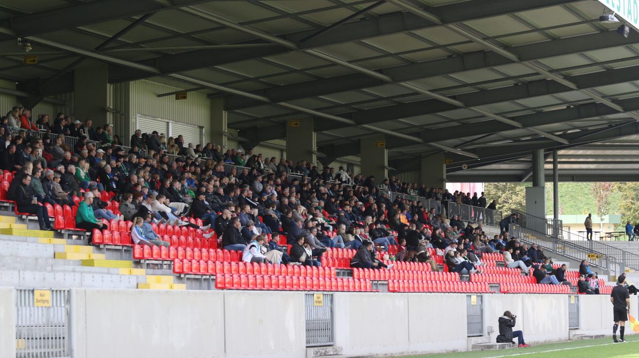Eine Menschenmenge sitzt in roten Sitzen in einem Stadion. Die Tribünen sind mit Zuschauern gefüllt, und einige stehen auf der Galerie. Das Stadion hat eine Metalldecke mit einem gelben Schild.