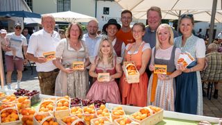 Mehrere Menschen posieren für ein Foto auf einem Freimarkt und halten Obst in den Händen. Sie tragen traditionelle bayerische Kleidung. Kisten mit Pfirsichen und Kirschen stehen vor ihnen auf dem Tisch.