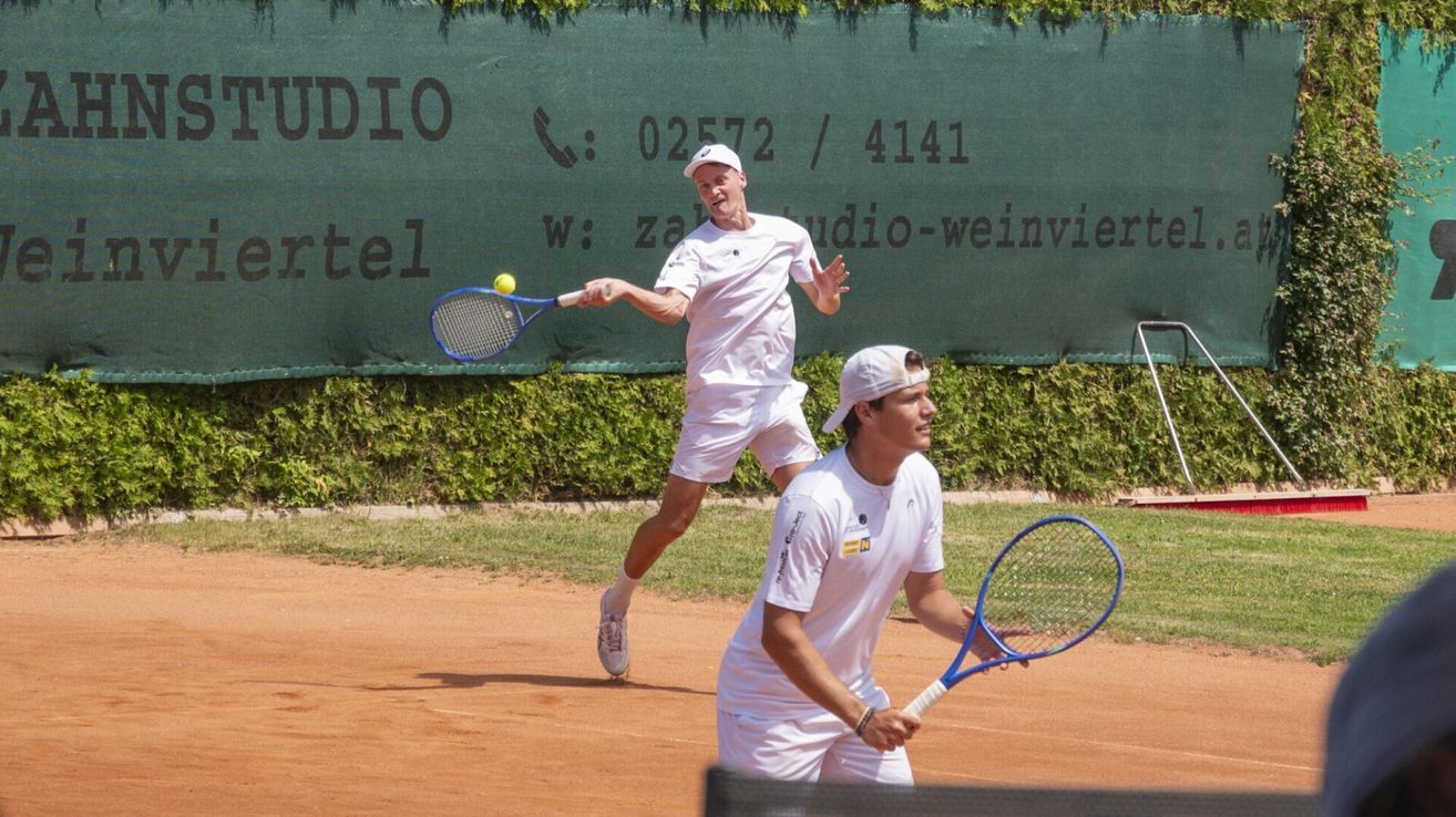 Zwei Männer spielen Tennis auf einem Sandplatz. Ein Mann springt und schlägt einen gelben Ball. Der andere Mann bereitet sich darauf vor, den Ball zu schlagen. Sie tragen beide weiße Uniformen.