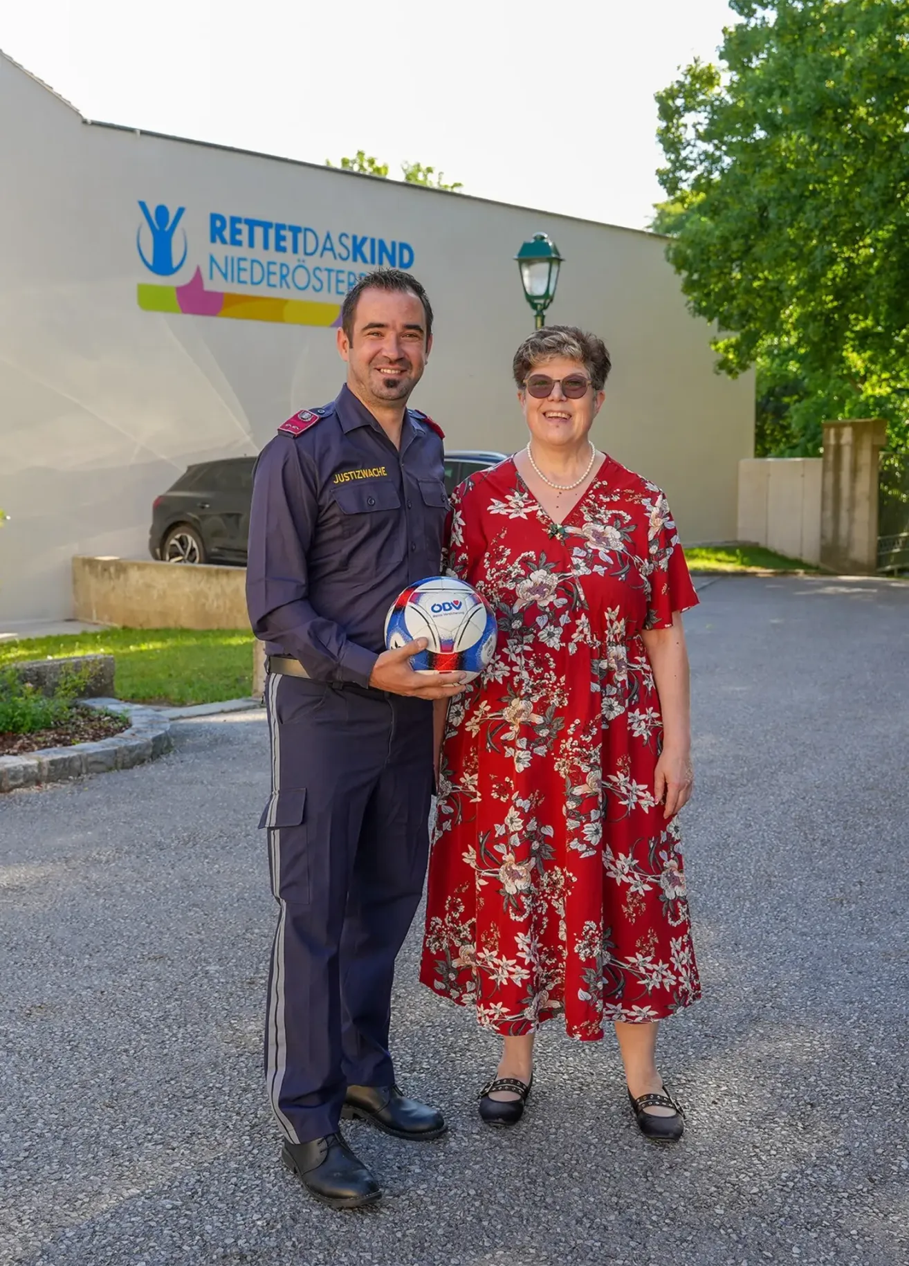 Ein Mann und eine Frau stehen vor einem Gebäude und lächeln. Der Mann hält einen Fußball, und beide sind formell gekleidet. Dahinter sind ein Auto und eine Straßenlaterne zu sehen.