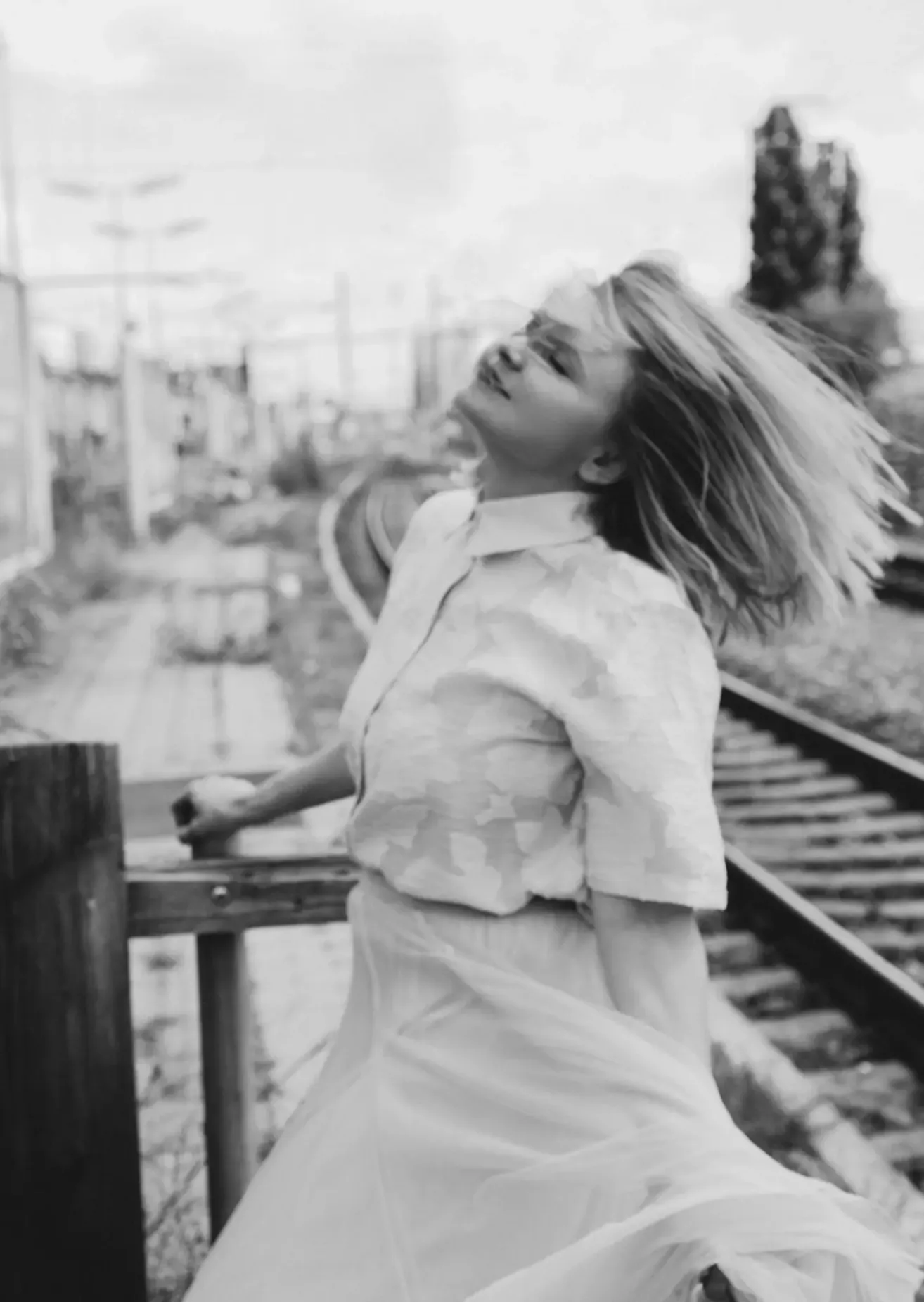 Eine Frau in einem weißen Kleid steht in der Nähe eines Zauns, ihr Haar weht im Wind neben den Bahngleisen.
