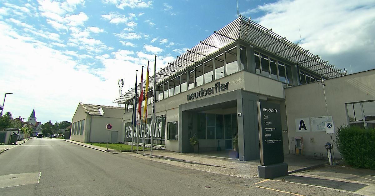 Ein modernes Gebäude namens 'neudoerfler' mit Fahnen und Topfpflanzen vor dem Eingang. Der Himmel ist klar mit einigen Wolken.