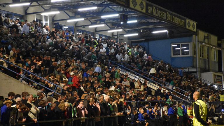 Eine große Menschenmenge versammelt sich in einem Stadion mit blauen Barrieren, beleuchtet von Deckenleuchten. Einige Fans tragen grüne und weiße Schals. Ein Banner mit der Aufschrift 'SV Klocher Bau'.