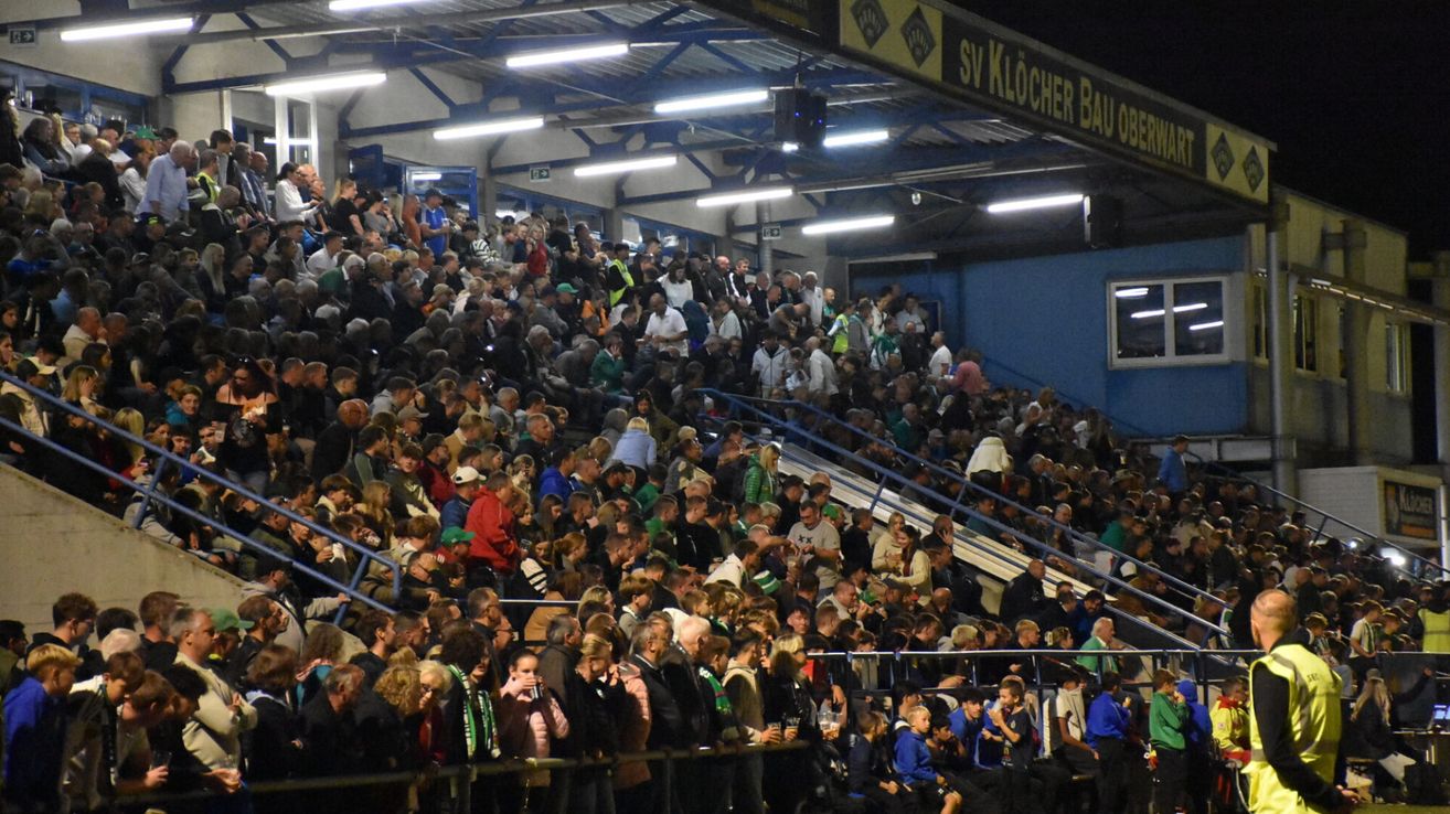 Eine große Menschenmenge versammelt sich in einem Stadion mit blauen Barrieren, beleuchtet von Deckenleuchten. Einige Fans tragen grüne und weiße Schals. Ein Banner mit der Aufschrift 'SV Klocher Bau'.