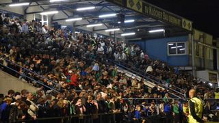 Eine große Menschenmenge versammelt sich in einem Stadion mit blauen Barrieren, beleuchtet von Deckenleuchten. Einige Fans tragen grüne und weiße Schals. Ein Banner mit der Aufschrift 'SV Klocher Bau'.