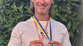 Ein lächelnder Mann mit langen Haaren hält zwei Medaillen vor einem grünen Buschhintergrund. Er trägt ein weißes Shirt und hat zwei Medaillen um den Hals.