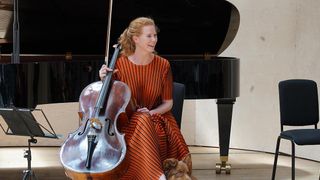 Eine Frau sitzt auf einem Stuhl, hält ein Cello und lächelt. Sie trägt ein orangefarbenes Kleid mit vertikalen Streifen. Hinter ihr steht ein schwarzer Flügel, der teilweise geöffnet ist. Ein Hund ruht neben ihr.