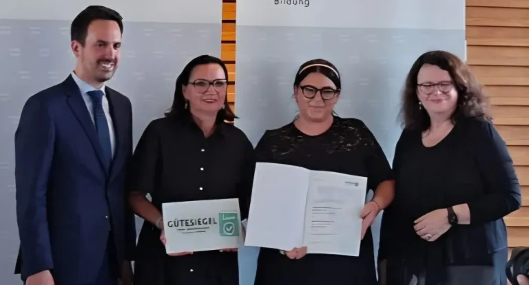 Zwei Frauen stehen zusammen, eine hält ein Zertifikat mit der Aufschrift 'Guteschein' und die andere ein offenes Dokument. Beide tragen Brillen. Dahinter ist eine Wand mit dem Wort 'Bildung'.