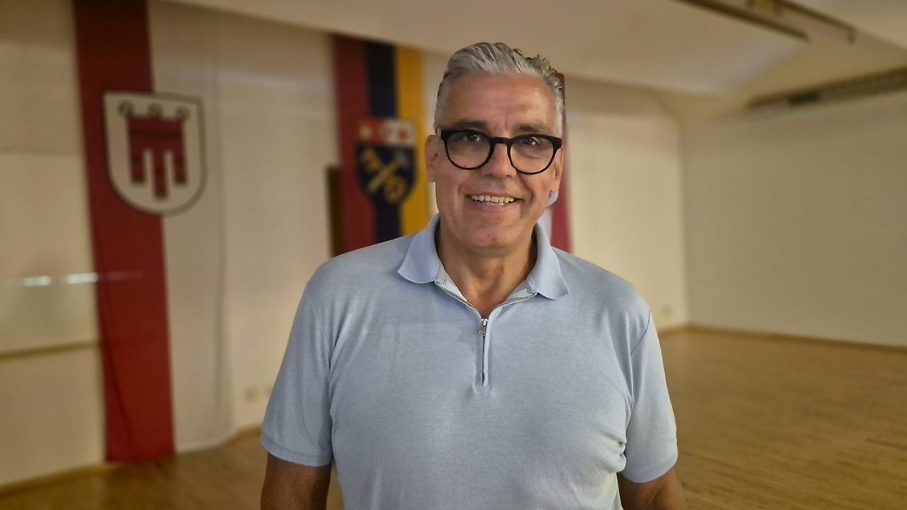 Ein Mann mit einer Brille und einem blauen Poloshirt steht in einem Raum mit Holzboden und einer Flagge dahinter.