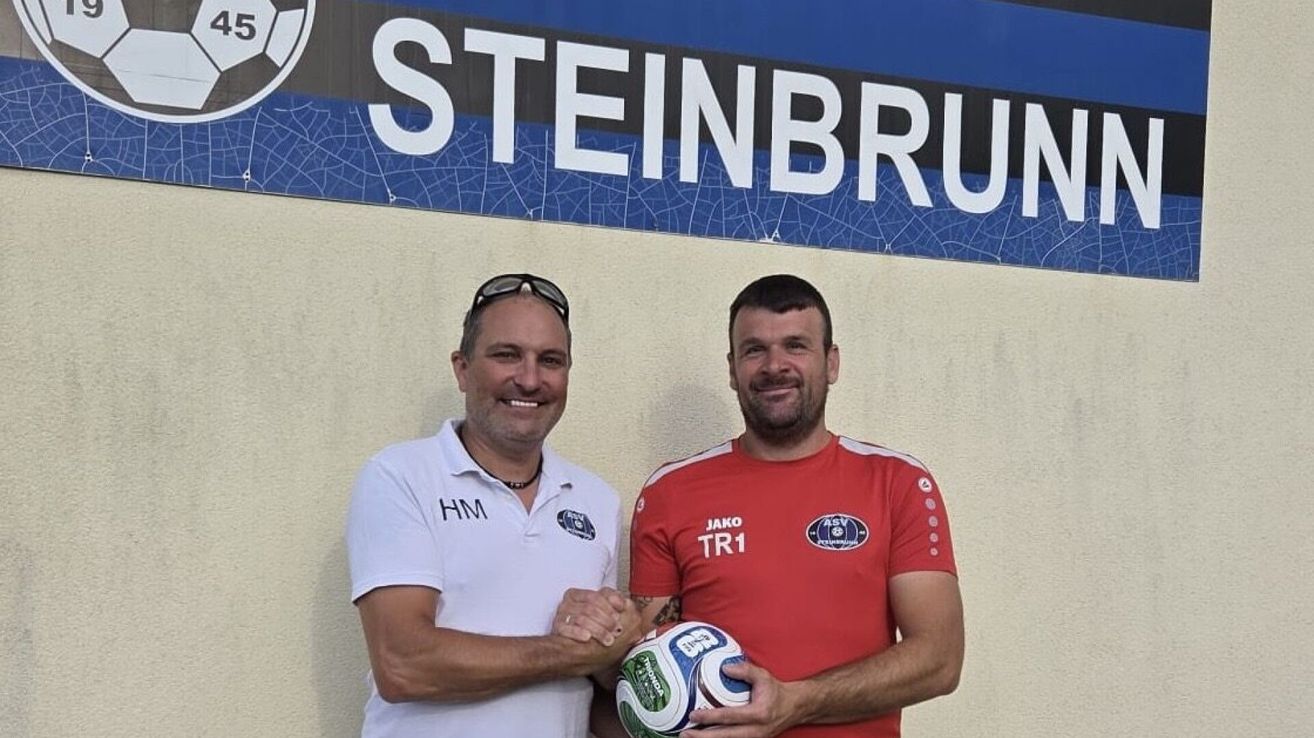 Zwei Männer, einer in einem weißen Poloshirt und der andere in einem roten Trikot, stehen nebeneinander, lächeln und halten einen Fußball vor einem Schild mit der Aufschrift STEINBRU.
