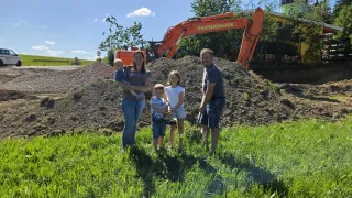 Eine vierköpfige Familie, einschließlich eines Babys, posiert auf einem Rasen mit einem großen orangefarbenen Bagger im Hintergrund.