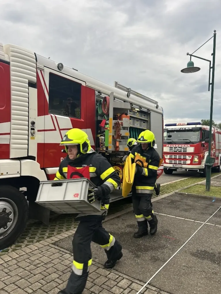 Two firefighters in full gear are unloading equipment from a fire truck on a paved road. Another fire truck is parked in the background.