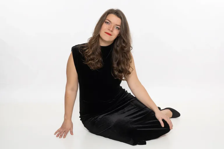 Eine Frau mit langen braunen Haaren, die ein schwarzes Kleid und Make-up trägt, sitzt auf einer weißen Oberfläche.