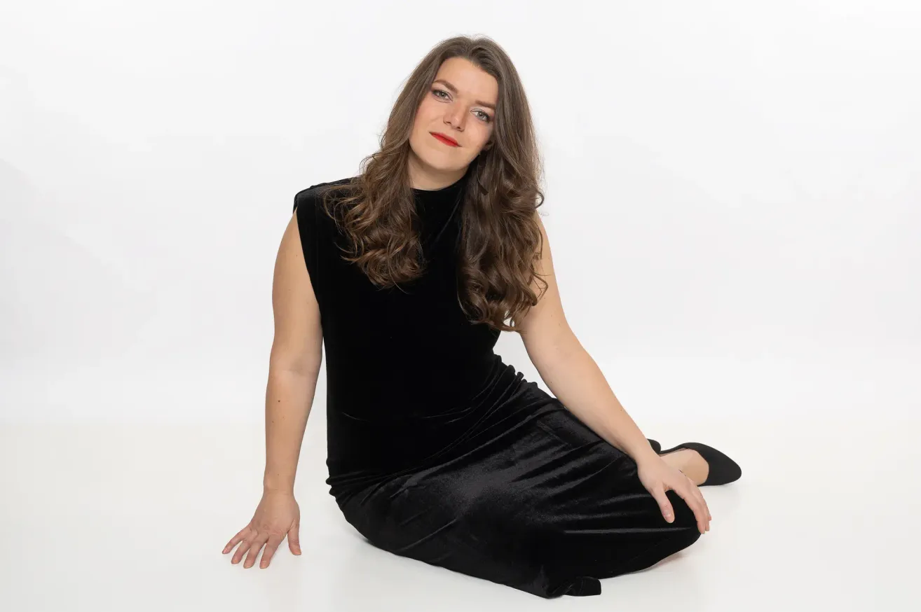 Eine Frau mit langen braunen Haaren, die ein schwarzes Kleid und Make-up trägt, sitzt auf einer weißen Oberfläche.