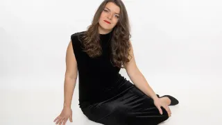 Eine Frau mit langen braunen Haaren, die ein schwarzes Kleid und Make-up trägt, sitzt auf einer weißen Oberfläche.