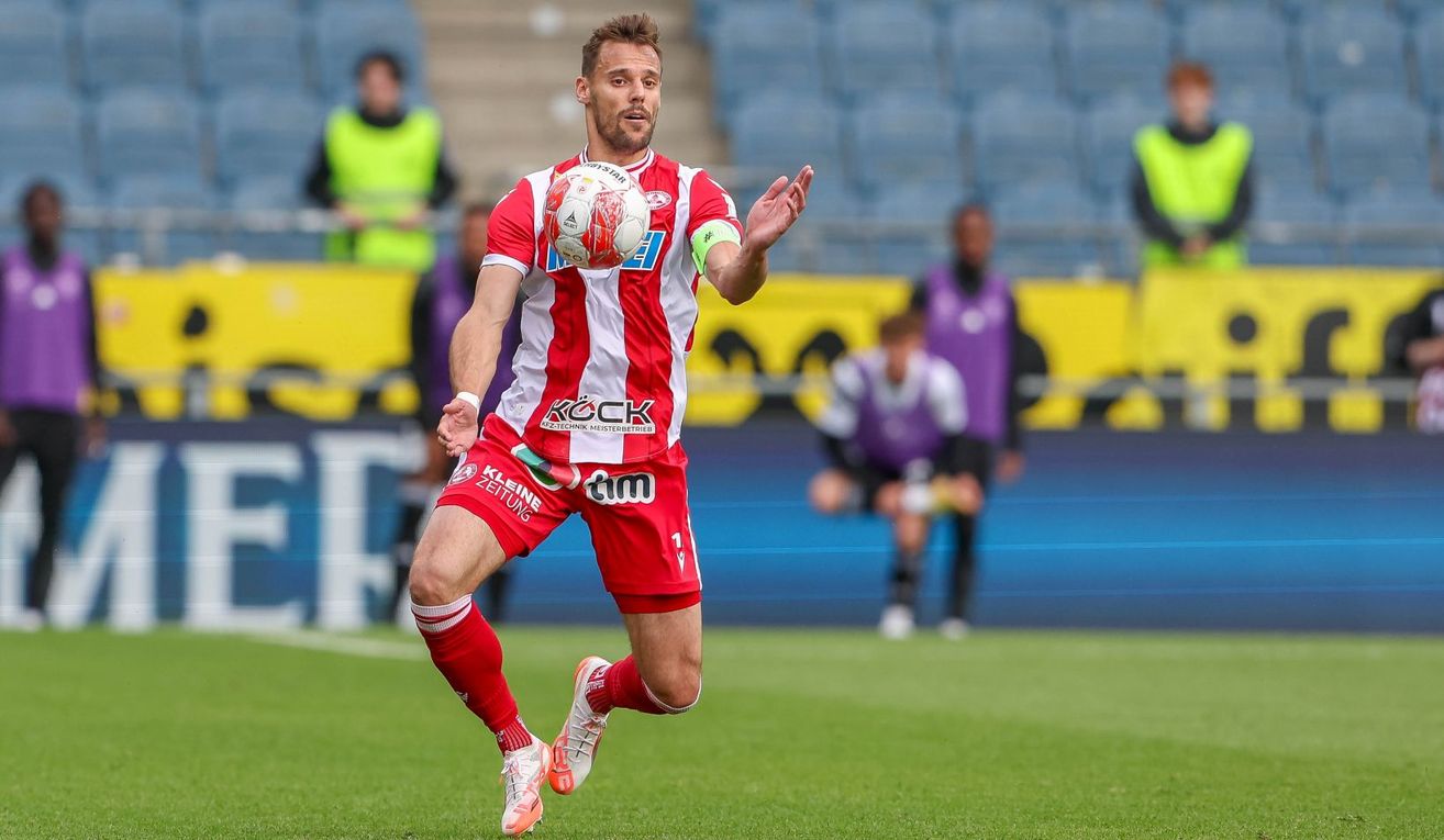 Ein Fußballspieler in einem roten Trikot läuft und tritt einen Ball. Er ist in der Mitte des Feldes, mit verschwommenen Personen im Hintergrund.
